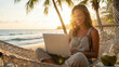 © Matias - Mujer joven y feliz trabajando como nómada digital con su laptop en una hamaca en la playa al atardecer.
