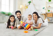 © Studio Romantic - Portrait of smiling family of four with toys on rug at home looking at camera. Happy parents, older daughter and baby child playing with colorful stacking rings and creating joyful memories together