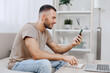 © SHOTPRIME STUDIO - Smiling man using a smartphone while sitting on a cozy sofa, creating a relaxed atmosphere with soft natural lighting and a minimalistic background.
