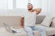 © SHOTPRIME STUDIO - Young man experiencing neck pain while working on a laptop, sitting comfortably on a couch. Light, airy, and modern home office setting enhances the atmosphere.