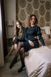 © svetograph - Women Fashion Dresses Bedroom: Indoor photoshoot showcasing two models in elegant black dresses, posing on a bed, showcasing autumn/winter fashion.