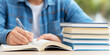 © Bonsales - Student hand writing notes in an open notebook with a pen, sitting at a desk with a stack of textbooks nearby