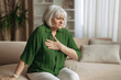 © Anastasiya - Senior caucasian woman leaning forward and touching chest at home, elderly female feeling shortness of breath or pain, healthcare and aging lifestyle concept indoors