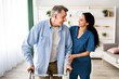 © Prostock-studio - A senior man is using a walker with assistance from a caregiver. They are both smiling in a well-lit room that has light-colored furniture. This moment shows warmth and care.