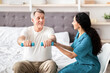 © Prostock-studio - A senior man is sitting on a bed holding small weights as a woman provides encouragement and assistance. They are in a bright room designed for physical activity.