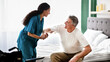 © Prostock-studio - A healthcare worker with curly hair helps an older man in a white shirt while he sits on the edge of a bed. They are engaged and smiling in a well-lit bedroom setting.