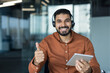 © Liubomir - Smiling male call center agent wearing a headset, holding a digital tablet, and giving a thumbs up gesture, signifying excellent customer service and approval in a modern office setting