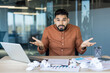 © Liubomir - Young office worker shrugs in frustration at a messy desk, laptop open amid crumpled papers, showing confusion, stress and uncertainty while facing a deadline and problem-solving