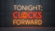 © Mr - Tonight: clocks forward, a bright neon sign on a dark textured wall announcing daylight saving