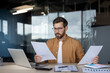 © Liubomir - Businessman focusing intently while analyzing paperwork and financial reports at his office desk, comparing information on documents with data displayed on a laptop screen