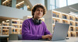 © umut hasanoglu - Young student sitting in library with laptop and headphones looking away thoughtfully