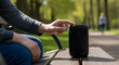 © Aura - Person interacting with a black wireless speaker device while sitting on a wooden bench in the sun