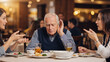© D-point - Older man with hearing aid struggling to hear women talking at a restaurant. Hearing loss concept affecting social interaction for senior.