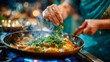 © Aliaksandra - Chef sprinkling fresh herbs into simmering curry sauce in a hot skillet over gas flame, steam rising, close-up cooking scene