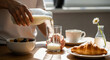 © Mahfud Picture - Close-up of hands pouring fresh milk from a bottle into a glass on a wooden table. A healthy morning breakfast is laid out with cereal, coffee, and a croissant