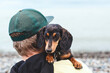 © Evgeniia Primavera - Cute dachshund dog resting on shoulder of man wearing green cap outdoors, small black tan puppy looking at camera, warm human animal bond, companionship travel lifestyle copyspace concept