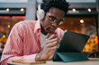 © BullRun - African freelancer with headphones drawing on tablet, deeply focused on digital task in cozy café setting, symbolizing tech-powered remote work and quiet concentration.