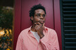© BullRun - Thoughtful African man in striped shirt touching chin, standing against red wall, showing deep reflection, critical thinking and self-awareness in urban creative setting.