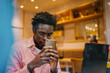 © BullRun - Young African man wearing glasses stares at smartphone through café glass, showing digital immersion, mobile tech focus and intense attention in remote freelancer lifestyle.