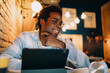 © BullRun - Smiling young man using tablet at cafe, illuminated by warm lighting, portraying modern connectivity, remote productivity, and positive engagement with digital tools in a creative workspace.