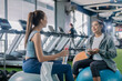 © whyframeshot - Two cheerful Asian women chatting and laughing together at a gym, one holding a smartphone while talking to her friend, enjoying a fun break between workouts.