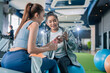 © whyframeshot - Two cheerful Asian women chatting and laughing together at a gym, one holding a smartphone while talking to her friend, enjoying a fun break between workouts.