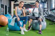 © whyframeshot - Two attractive Asian women resting together at a modern gym, sitting on exercise balls with water bottles, wearing colorful sportswear in a bright fitness club.