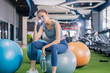 © whyframeshot - confident Asian woman sitting on exercise balls at a modern gym, wearing a gray sports top and blue leggings with white sneakers, posing with a calm and focused look.