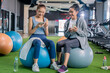 © whyframeshot - Two cheerful Asian women chatting and laughing together at a gym, one holding a smartphone while talking to her friend, enjoying a fun break between workouts.