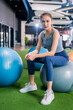 © whyframeshot - confident Asian woman sitting on exercise balls at a modern gym, wearing a gray sports top and blue leggings with white sneakers, posing with a calm and focused look.