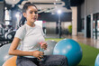 © whyframeshot - smiling Asian woman resting at the gym, holding a water bottle while sitting on an exercise ball, wearingactive sport cloth leggings in a modern fitness club.