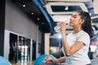 © whyframeshot - smiling Asian woman resting at the gym, holding a water bottle while sitting on an exercise ball, wearingactive sport cloth leggings in a modern fitness club.