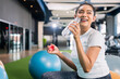 © whyframeshot - smiling Asian woman resting at the gym, holding a water bottle while sitting on an exercise ball, wearingactive sport cloth leggings in a modern fitness club.