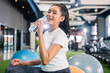 © whyframeshot - smiling Asian woman resting at the gym, holding a water bottle while sitting on an exercise ball, wearingactive sport cloth leggings in a modern fitness club.