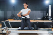 © whyframeshot - Asian woman doing Pilates reformer exercise in a studio, kneeling and pulling resistance band with focused concentration, wearing white top and black leggings.