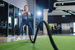 © whyframeshot - fit Asian woman doing battle rope exercise at a modern gym, holding thick black ropes in a wide stance, wearing sport active cloth leggings with intense focus.