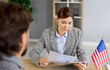 © Studio Romantic - Consular visa interview and immigrants documents discussion of female lawyer and man at office desk with US flag. USA immigration application and paperwork review at meeting by embassy employee