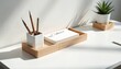© Vadym - Minimalist desk setup with wooden tray organizer holding white pen cup, notepad, and pencils. A green plant in white pot sits nearby. Sunlight creates shadows on the white surface.