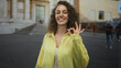© Krakenimages.com - Young woman smiling while pointing with right hand and making ok sign with left hand in front of a building on a city street; joy greeting optimism.