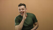 © Krakenimages.com - Young man whispering with a playful expression in front of a plain brown background, wearing a green shirt and showing tattoos on his arms, conveying secrecy and intrigue.