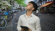 © Krakenimages.com - Young man in white shirt holding tablet looking up on a busy city street near bicycles and cafe seating in daylight; curiosity planning concentration.