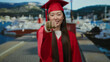 © Krakenimages.com - Woman in red graduation gown celebrates at seaside port with boats visible in the background under bright sunlight, showcasing an inspiring and joyful moment.