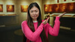 © Krakenimages.com - Woman wearing pink sweater gesturing in an art gallery with a thoughtful expression against a backdrop of paintings displaying an interior museum setting with warm lighting.