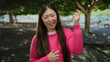 © Krakenimages.com - Woman in a pink sweater gestures playfully outdoors in a park with trees, smiling and enjoying a sunny day.