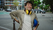 © Krakenimages.com - Hispanic man with headphones and hardhat stands on street clutching his back, showing discomfort while wearing casual clothing in an urban outdoor setting.