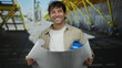 © Krakenimages.com - Hispanic man wearing hardhat smiles while holding blueprints on bright outdoor street.