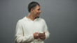 © Krakenimages.com - Man holding a smartphone, hands texting and glancing sideways in a gray studio while wearing white sweater and gold chain; contemplative focus.