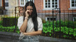 © Krakenimages.com - Young hispanic woman wiping tears with hand, arm crossed, standing by iron fence in front of building; sadness vulnerability isolation.