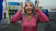 © Krakenimages.com - Senior woman with grey hair smiling playfully outdoors at a petrol station, spontaneously expressing joy while covering and uncovering her eyes joyfully.