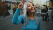 © Krakenimages.com - Woman saluting with fingers on head, grey-haired, wears blue sweater, standing on outdoor terrace of busy street restaurant showing mature elegance and cheerful expression.